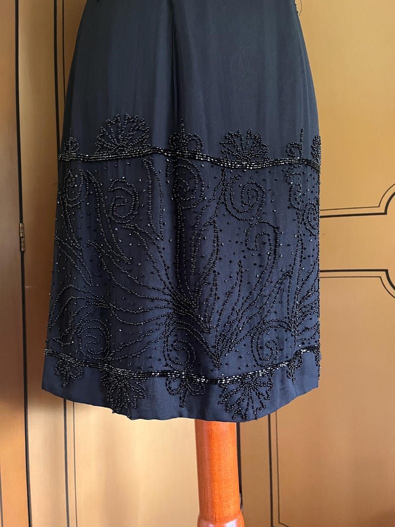 May include: A dark navy blue dress with intricate beaded embellishments. The dress features a detailed pattern of swirling designs and floral motifs along the hemline. The beading adds texture and visual interest to the elegant garment.