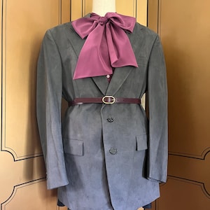 Pu&ograve; includere: Un blazer grigio con un papillon e una cintura bordeaux, esposto su un manichino. Il blazer ha una chiusura a due bottoni e tasche anteriori. La cintura ha una fibbia color oro.