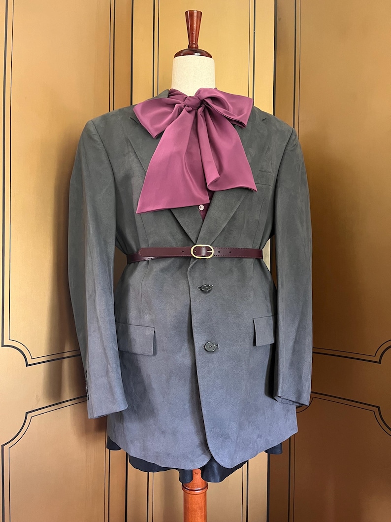 Pu&ograve; includere: Un blazer grigio con un papillon e una cintura bordeaux, esposto su un manichino. Il blazer ha una chiusura a due bottoni e tasche anteriori. La cintura ha una fibbia color oro.