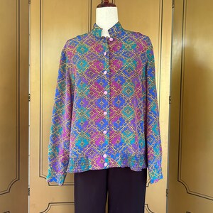 May include: A long-sleeved button-up shirt with a colorful geometric pattern in shades of blue, purple, green, and gold. The shirt has a mandarin collar and a pink button closure.