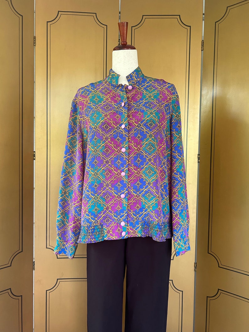 May include: A long-sleeved button-up shirt with a colorful geometric pattern in shades of blue, purple, green, and gold. The shirt has a mandarin collar and a pink button closure.