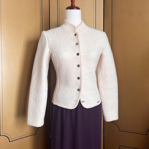 Op de afbeelding: Cr&egrave;mekleurige jas met een hoge kraag en donkere knopen. De jas wordt gepresenteerd op een mannequin, gecombineerd met een lange, donkerpaarse rok. De jas heeft een getailleerde pasvorm en een getextureerde uitstraling.