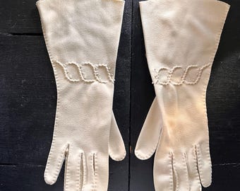 Könnte beinhalten: Ein Paar cremefarbene Handschuhe mit einem dekorativen, wellenförmigen Design am oberen Rand. Die Handschuhe sind aus einem weichen, strukturierten Material gefertigt und haben einen klassischen, eleganten Stil. Die Handschuhe liegen auf einer dunklen Oberfläche.