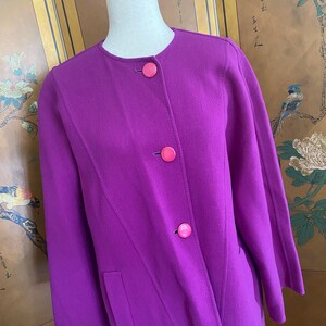 May include: A vibrant purple coat with a round neckline and three pink buttons. The coat features a tailored design with a diagonal seam and two front pockets. The sleeves are three-quarter length.