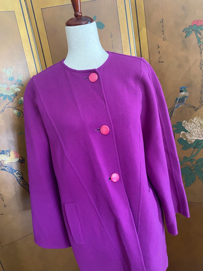 May include: A vibrant purple coat with a round neckline and three pink buttons. The coat features a tailored design with a diagonal seam and two front pockets. The sleeves are three-quarter length.