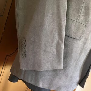 Pu&ograve; includere: Primo piano di un blazer grigio con tre bottoni coordinati sulla manica. Il blazer ha un aspetto strutturato, simile alla pelle scamosciata e una singola tasca. L'indumento &egrave; esposto su un manichino.