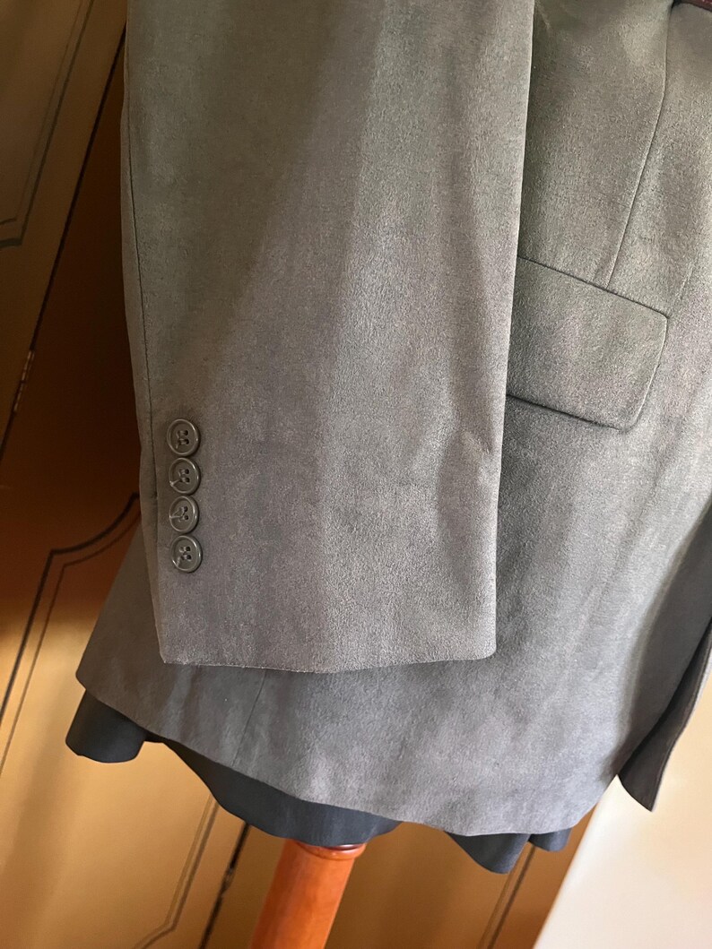 Pu&ograve; includere: Primo piano di un blazer grigio con tre bottoni coordinati sulla manica. Il blazer ha un aspetto strutturato, simile alla pelle scamosciata e una singola tasca. L'indumento &egrave; esposto su un manichino.