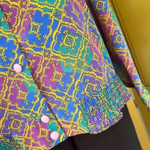May include: A purple, blue, and green patterned blouse with a yellow geometric design. The blouse has a ruffled hem and pink buttons.
