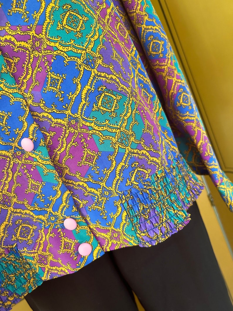 May include: A purple, blue, and green patterned blouse with a yellow geometric design. The blouse has a ruffled hem and pink buttons.