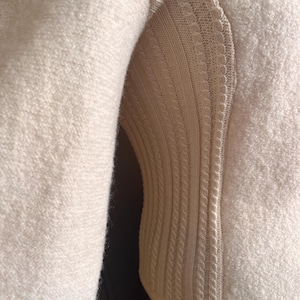 Op de afbeelding: Close-up van een cr&egrave;mekleurige trui. De mouw bevindt zich aan de linkerkant, met een getextureerde, fuzzy stof. De rechterkant heeft een gebreid paneel met een kabelbreipatroon en decoratieve randen. De trui lijkt gemaakt van een zacht, warm materiaal.