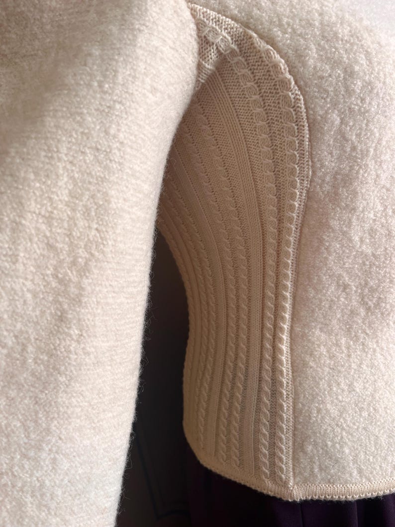 Op de afbeelding: Close-up van een cr&egrave;mekleurige trui. De mouw bevindt zich aan de linkerkant, met een getextureerde, fuzzy stof. De rechterkant heeft een gebreid paneel met een kabelbreipatroon en decoratieve randen. De trui lijkt gemaakt van een zacht, warm materiaal.