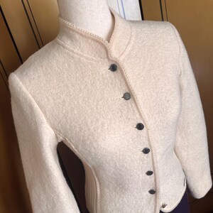 Op de afbeelding: Cr&egrave;mekleurige wollen jas met een opstaande kraag en een rij zwarte knopen aan de voorkant. De jas heeft lange mouwen en een getailleerde pasvorm, met decoratieve stiksels langs de randen. De jas wordt op een mannequin getoond.