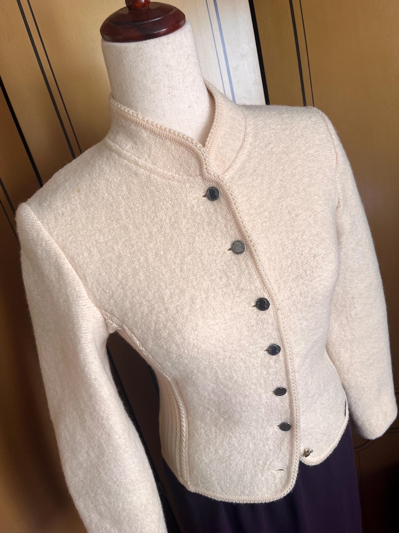 Op de afbeelding: Cr&egrave;mekleurige wollen jas met een opstaande kraag en een rij zwarte knopen aan de voorkant. De jas heeft lange mouwen en een getailleerde pasvorm, met decoratieve stiksels langs de randen. De jas wordt op een mannequin getoond.
