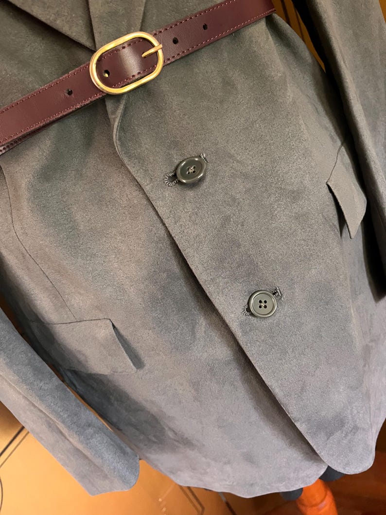 Pu&ograve; includere: Un blazer grigio con due bottoni e una cintura bordeaux con fibbia dorata. Il blazer ha due tasche e una morbida texture simile alla pelle scamosciata. La cintura &egrave; stretta in vita.