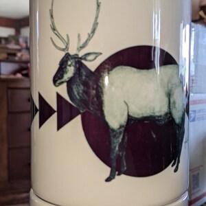 May include: White ceramic mug featuring a detailed illustration of an elk inside a dark burgundy circle. The elk is depicted in shades of gray and black, with large antlers. Geometric arrow shapes are on the left side of the mug.