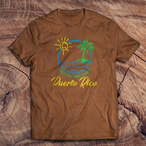 May include: A brown t-shirt with a graphic design of a sun, palm tree, and waves. The text "Isla del Encanto Puerto Rico" is printed in yellow on the shirt.