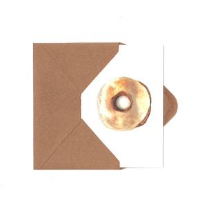 May include: A watercolor illustration of a donut on a white card, partially inserted into a brown envelope. The donut has a light brown exterior with a white center. The envelope is a solid brown color.