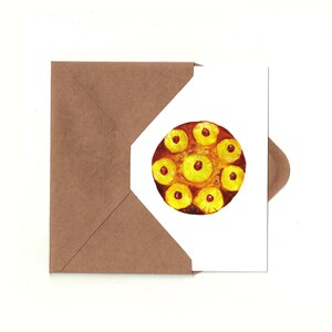 May include: A white card with a brown envelope partially covering it. The card has a watercolor painting of a pineapple upside-down cake with a brown crust and yellow pineapple slices topped with red cherries.