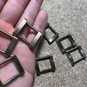 Puede incluir: Primer plano de varias hebillas metálicas cuadradas pequeñas con acabado de latón. Cada hebilla tiene un marco rectangular con una barra que atraviesa el centro. Las hebillas están dispersas sobre una superficie marrón claro.