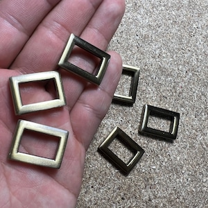 May include: A collection of antique brass-coloured rectangular belt buckles with a diamond shape. The buckles have a hollow centre and are made of sturdy metal. The buckles are shown on a textured surface.