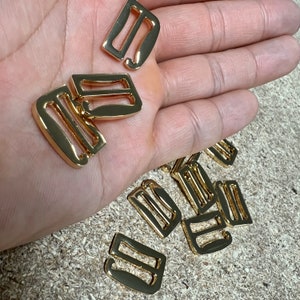 Op de afbeelding: Een verzameling goudkleurige metalen bh-bandjes schuifjes en haakjes. De hardware-onderdelen hebben verschillende vormen, waaronder rechthoekige schuifjes en gebogen haakjes. De items worden gepresenteerd op een lichtbruin oppervlak.