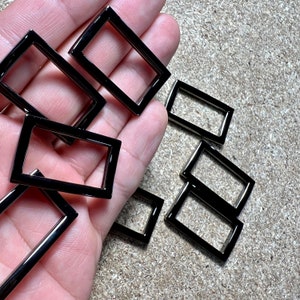 May include: Several rectangular, black metal frames with a glossy finish. The frames are held in a hand, and are scattered on a light brown surface. These shiny frames are likely for crafting or jewellery making. Each frame is a rectangle.