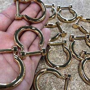 May include: A collection of shiny, gold-coloured metal D-ring buckles. Each buckle has a rounded D-shape with a straight bar that screws into the D-ring. These buckles are likely for use in leathercraft, bag making, or other crafting projects.