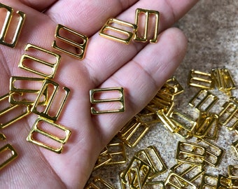 May include: A selection of small, shiny gold-coloured metal bra strap adjusters. The adjusters are rectangular with two parallel slots for the strap, and a hook on one side.