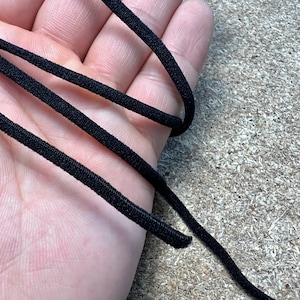 Puede incluir: Primer plano de varios hilos de cordón elástico negro. El cordón está tejido apretado y parece estar hecho de un material sintético. El cordón se sostiene en una mano, con el fondo desenfocado.