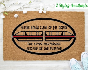 May include: A brown coir doormat with the text "PLEASE STAND CLEAR OF THE DOORS" and "POR FAVOR MANTENGASE ALEJADO DE LAS PUERTAS" in black text. A black and red silhouette of a monorail train is in the center of the doormat.