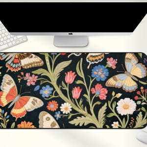 May include: A rectangular black mousepad featuring a colorful floral and butterfly design. The design includes butterflies in shades of orange, yellow, and blue, along with pink, blue, and white flowers. The mousepad is placed under a computer monitor and keyboard.
