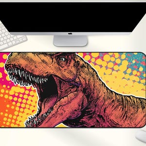 May include: A rectangular mousepad featuring a pop art style illustration of a Tyrannosaurus Rex with its mouth open. The background has a pink, yellow, and blue dot pattern. A computer monitor, keyboard, and mouse are visible.