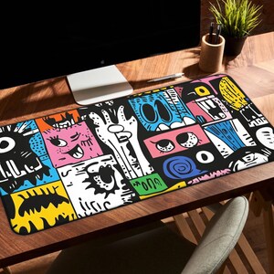 May include: A rectangular desk mat featuring a vibrant, multi-colored graphic print with various abstract cartoon-style faces, eyes, and shapes in black, white, pink, blue, yellow, and green, arranged in a dense, comic-inspired collage pattern.