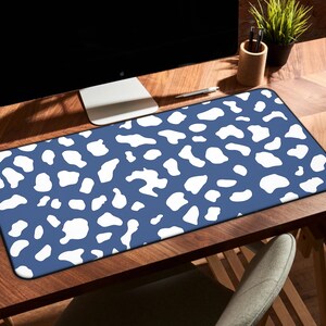 Puede incluir: Alfombrilla de escritorio rectangular con fondo azul y formas abstractas blancas. La alfombrilla está sobre un escritorio de madera, con un monitor de ordenador, un portalápices y una planta en el fondo. La alfombrilla mide unos 76 cm de largo.