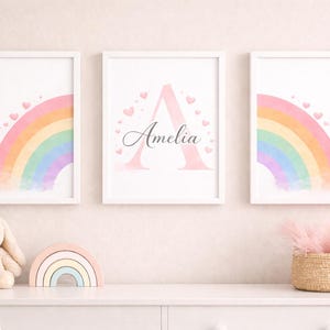 May include: Three framed prints with pastel rainbow designs. The central print features the name "Amelia" with a large letter "A" and heart accents. A plush bunny, a wooden rainbow, and a woven basket are on the shelf below.