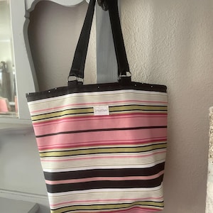 May include: A striped tote bag with black straps hangs from a hook. The bag features horizontal stripes in pink, green, brown, and white. A small white tag with the text "OrheartStudio?" is visible on the front.
