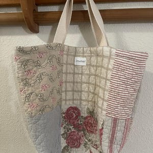 May include: A patchwork tote bag with a variety of quilted fabrics. The bag features floral, striped, and plaid patterns in shades of beige, pink, and red. The bag has cream-colored handles and a small tag that reads "OffycotStudio?".