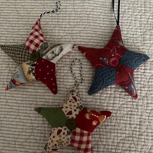 May include: Three stuffed fabric star ornaments featuring patchwork designs in red, green, blue, and white patterns. Each star is accented with a central button and includes a hanging loop made of either black and white twine or a solid navy ribbon.