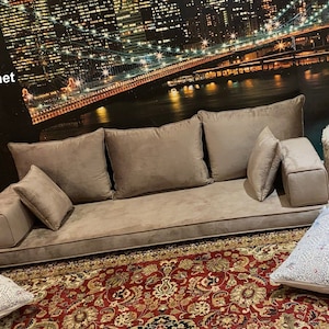 Op de afbeelding: Een grote, bruine vloersofa met een skyline van de stad als achtergrond. De bank heeft een dik, gestoffeerd kussen en meerdere kussens. De bank staat op een rood en gouden tapijt.