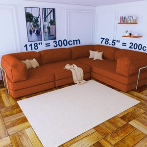 Pu&ograve; includere: Un grande divano angolare a forma di L in tessuto testurizzato color arancio bruciato con dettagli della struttura in metallo. Il divano misura 300 cm per 200 cm. Include diversi piccoli cuscini beige e una coperta beige drappeggiata.