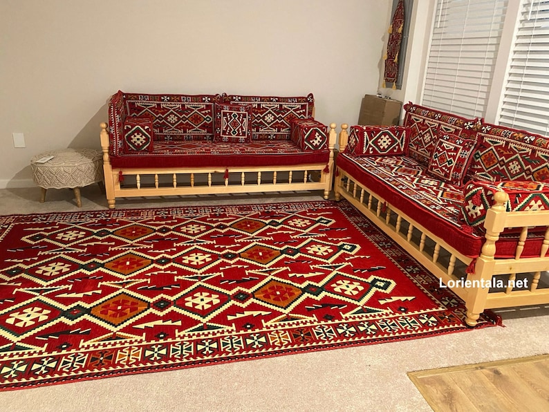 May include: Two wooden couches with red and white patterned cushions and a red patterned rug. The couches are made of light wood and have a simple design. The rug is a traditional Moroccan style with geometric patterns.