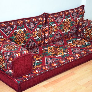 May include: A red floor cushion with a geometric pattern in shades of red, orange, green, and white. The cushion is made up of multiple pieces, including a large base cushion and several smaller cushions. The cushion is perfect for sitting on the floor or using as a sofa.