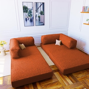Pu&ograve; includere: Due divani modulari da pavimento rivestiti in un tessuto a coste color arancio ruggine testurizzato. Ogni divano presenta una base rettangolare a profilo basso, un singolo cuscino per lo schienale attaccato e un piccolo cuscino quadrato chiaro.