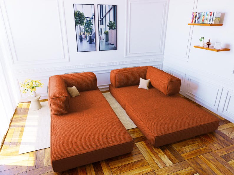 Pu&ograve; includere: Due divani modulari da pavimento rivestiti in un tessuto a coste color arancio ruggine testurizzato. Ogni divano presenta una base rettangolare a profilo basso, un singolo cuscino per lo schienale attaccato e un piccolo cuscino quadrato chiaro.