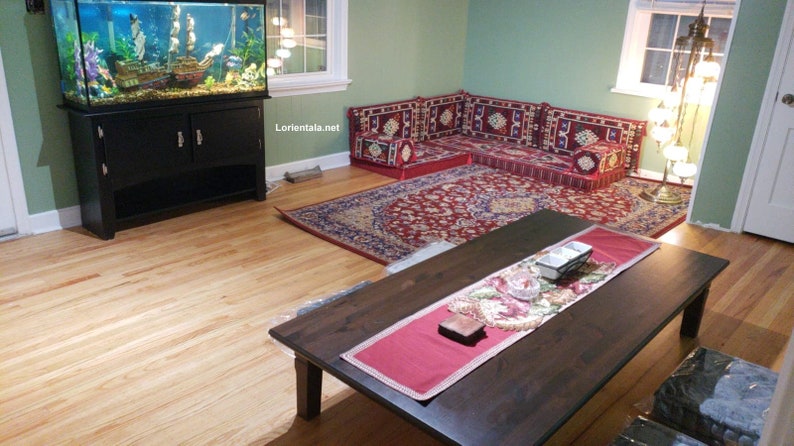May include: A living room with a large fish tank, a dark wood coffee table, and a red and blue patterned rug. The room is decorated with a traditional Middle Eastern style seating arrangement, featuring colorful cushions and pillows.