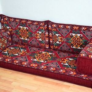 May include: A red and burgundy floor cushion with a geometric pattern in shades of green, yellow, and white. The cushion is made up of multiple pieces, including a large rectangular base and several smaller square cushions.