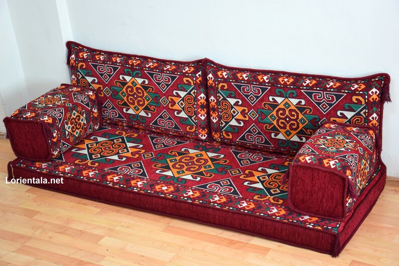 May include: A red and burgundy floor cushion with a geometric pattern in shades of green, yellow, and white. The cushion is made up of multiple pieces, including a large rectangular base and several smaller square cushions.