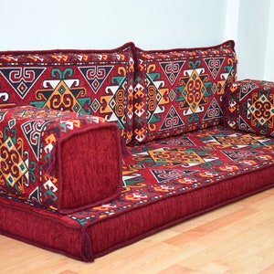 May include: A red floor cushion with a geometric patterned fabric. The cushion is made up of multiple pieces, including a large rectangular base and several smaller square cushions. The fabric features a mix of red, green, yellow, and white colors.