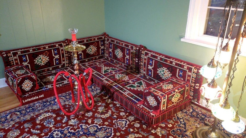 May include: A red and blue patterned couch with a matching rug and a red hookah on the floor. The couch is made up of individual cushions and is arranged in a traditional Moroccan style. A decorative lamp with multiple glass globes hangs in the background.