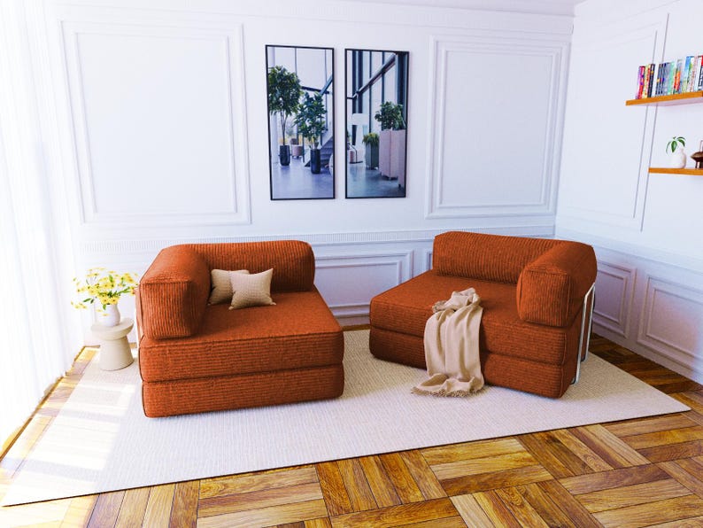 Pu&ograve; includere: Due poltrone modulari senza braccioli rivestite in un tessuto testurizzato color arancio bruciato. Ogni poltrona presenta un design basso con un solo bracciolo laterale, seduta quadrata e schienale. Una include due piccoli cuscini beige, l'altra un plaid beige.